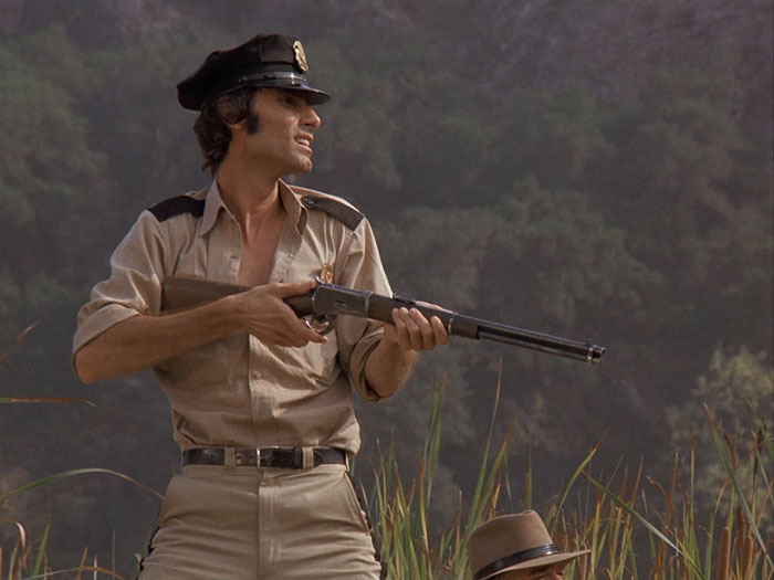 Karl [Anthony James] in guard uniform holding a rifle and standing on top of the patrol car (not shown)