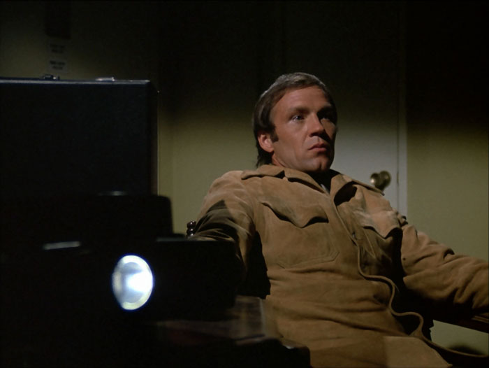 John Horn as the hitman in brown suede jacket, sitting expressionless, watching his slide show