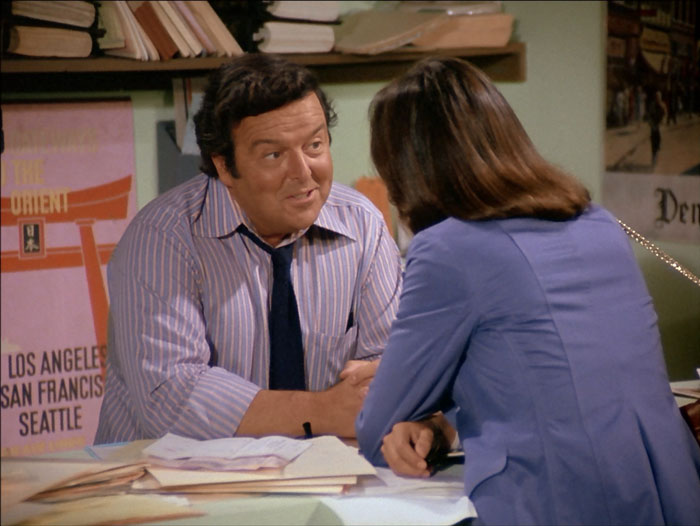 David Healy as Miles Cavendish, behind the counter in his travel agency with Sabrina leaning over the counter