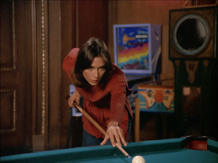 Sabrina lines up her pool shot. She's wearing an orange brown shirt and jeans. There are colorful pinball machines in the background.