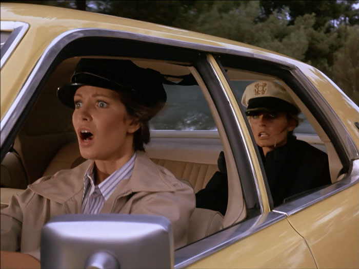 Kelly driving a cab with Jill in the back seat. Kelly is wearing a cabbie hat. Jill is wearing a captain's hat. Both have horrified looks on their faces.