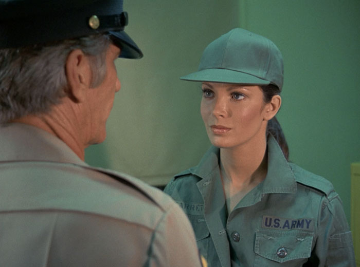 Over the shoulder shot on Kelly (Jaclyn Smith) in army fatigues and cap talking to Billings (L.Q. Jones) The predominant color in the shot is green.