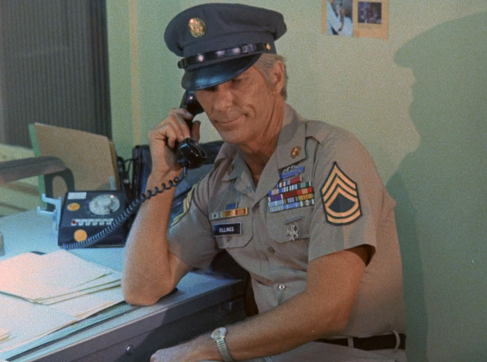 Billings (L.Q. Jones) in uniform, sitting at his desk talking on the phone.