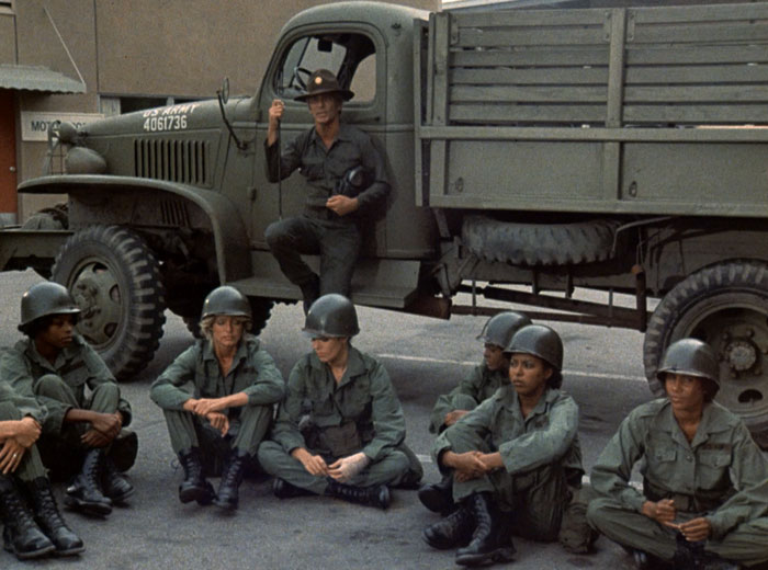 Billings (L.Q. Jones) leans against an army truck in the background and the recruits sit on the ground in front of him.