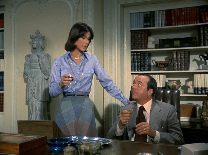 Sabrina (Kate Jackson) stands next to Bosley at his desk. She is wearing a silky, light blue blouse with a patterned skirt and thin belt.